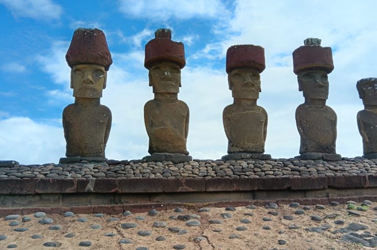 Five Moai statues with red topknots against a blue sky.